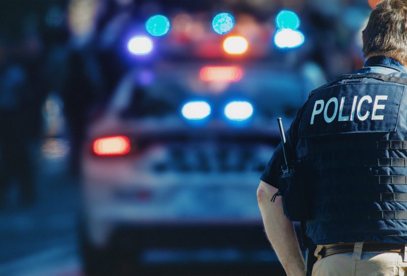 American,Policeman,And,Police,Car,In,The,Background